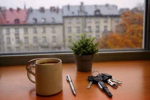 Biurko przy oknie z kubkiem kawy i kluczami, za szybą kamienice Wrocławia w deszczowy poranek